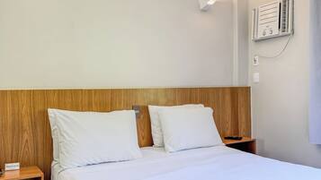 Habitación de hotel con cabecera de madera, ropa de cama blanca, mesita de noche con lámpara y aire acondicionado de pared.