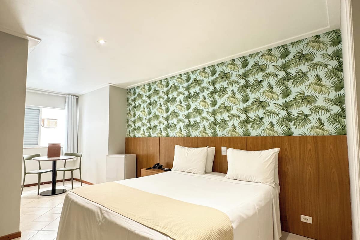 Habitación de hotel con una cama grande, cabecera de madera, una mesita con sillas y una ventana con cortinas.