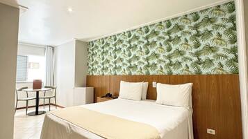 Habitación de hotel con una cama grande, cabecera de madera, una mesita con sillas y una ventana con cortinas.