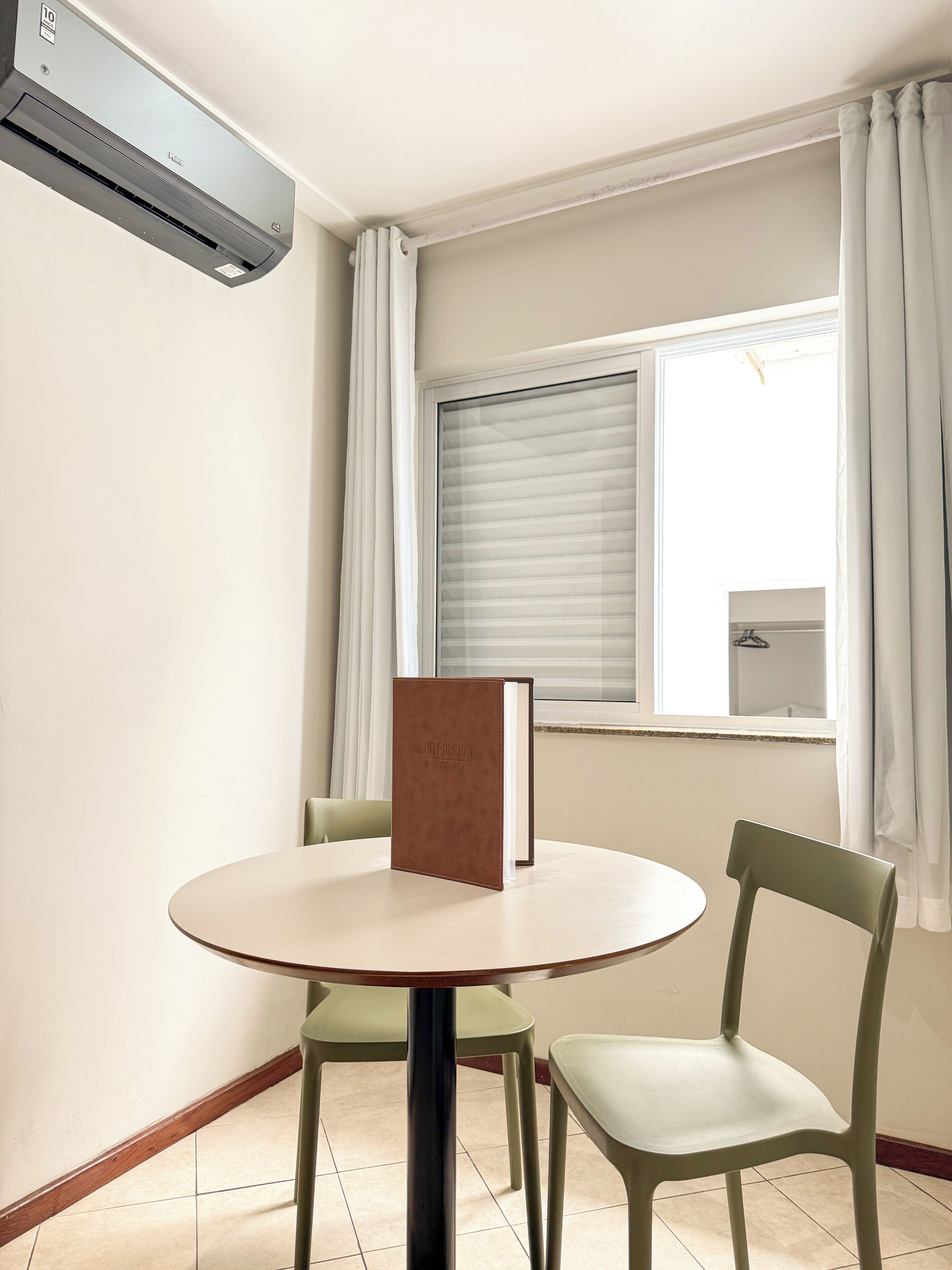 Ein Zimmer mit einem runden Tisch, zwei Stühlen, einem Fenster mit Jalousien und einer Klimaanlage.