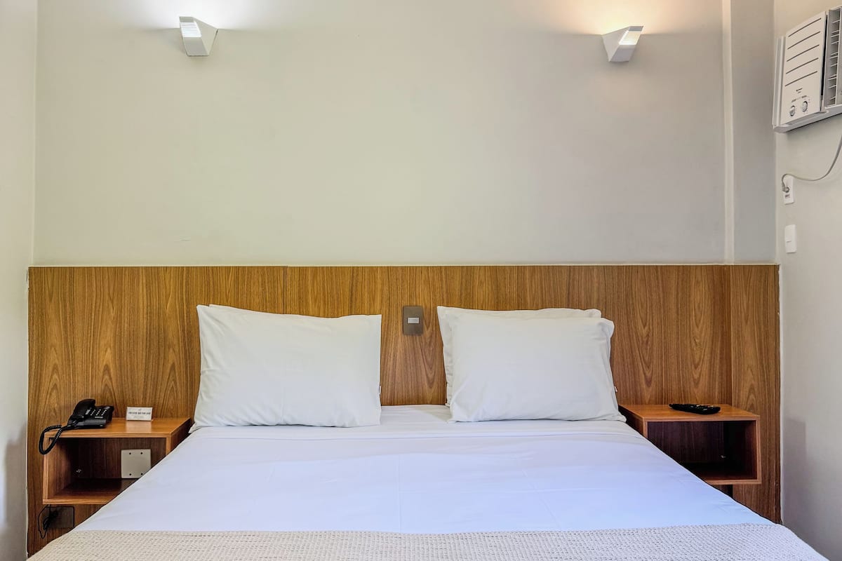 Habitación de hotel con cabecera de madera, dos mesitas de noche, cama con ropa blanca y dos lámparas fijadas a la pared.