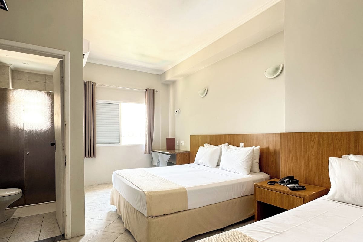 Habitación de hotel con dos camas, cabecero de madera, mesita de noche con teléfono y control remoto, y baño visible a través de una puerta abierta.