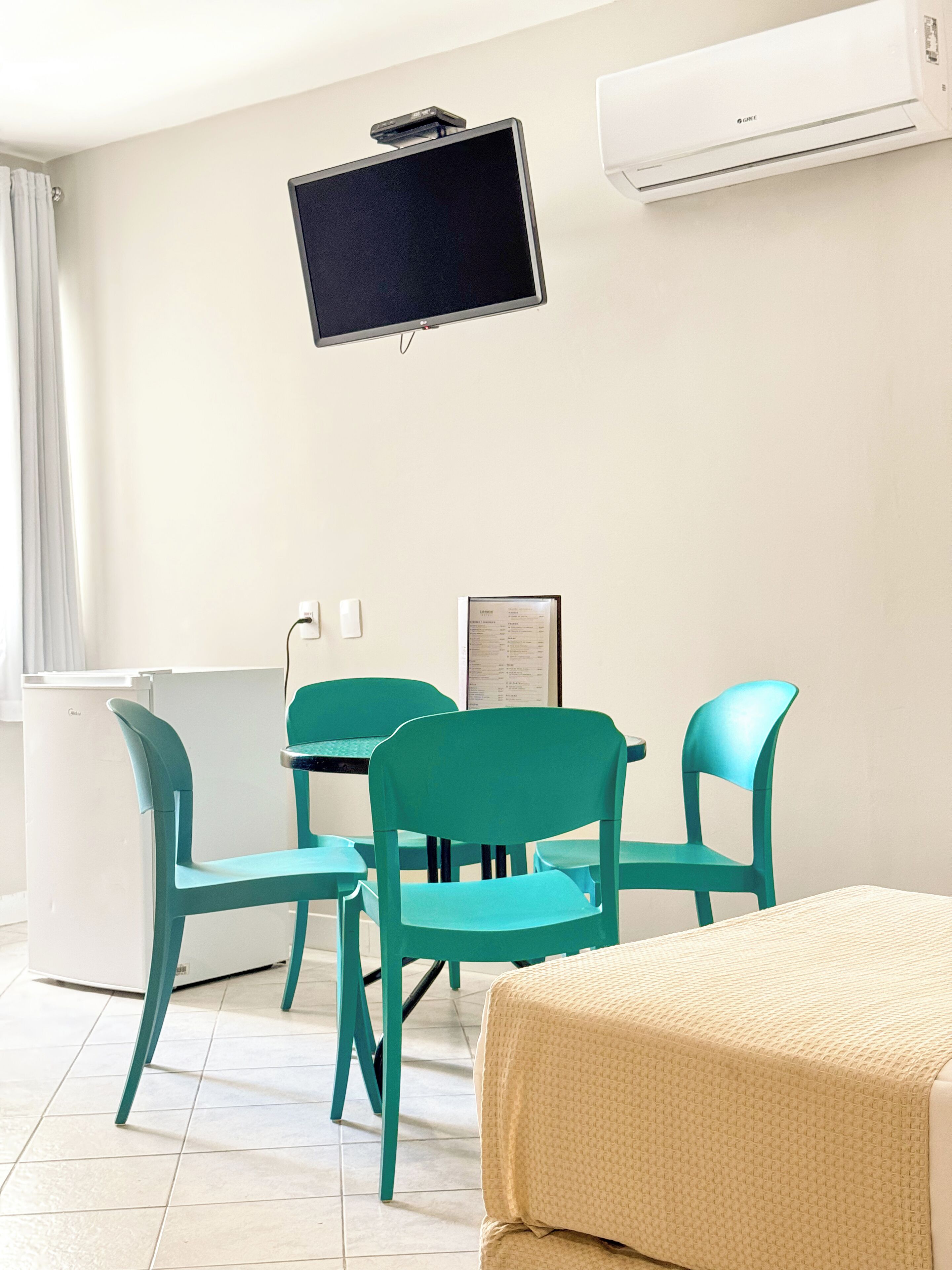 A room with teal chairs, a white bed, a flat-screen TV, and an air conditioning unit.