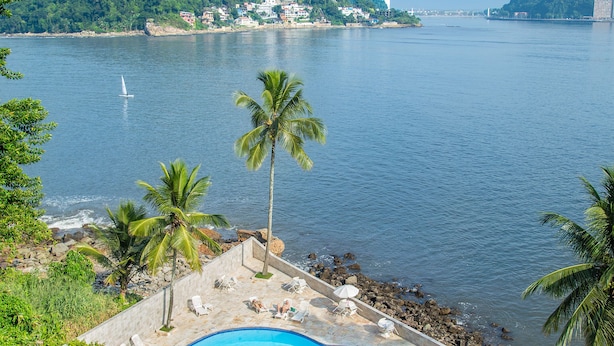 Una piscina con sillones de descanso y palmeras, rodeada por una terraza de piedra y con vista a un cuerpo de agua donde se ven edificios y colinas a lo lejos.