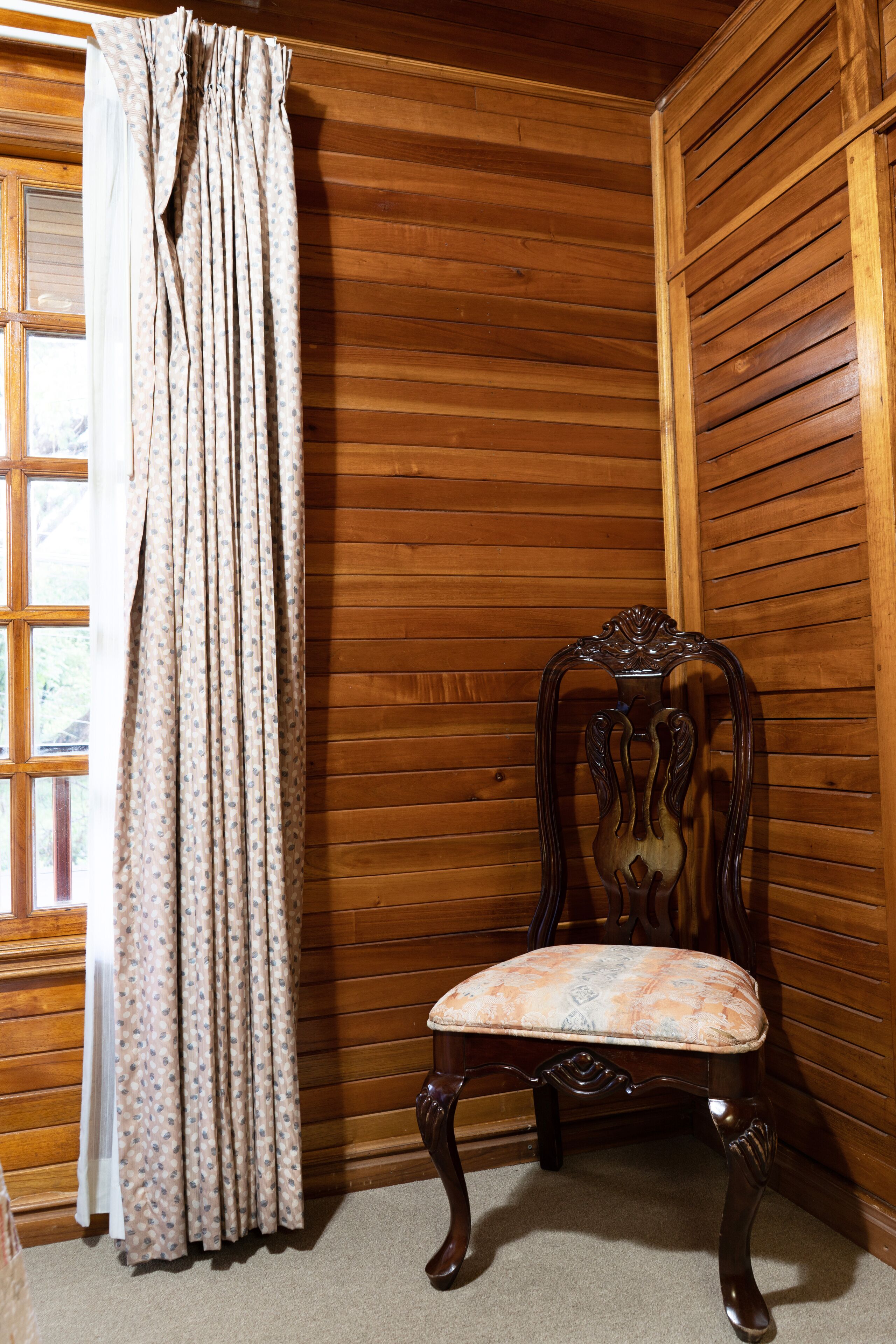 A corner room with wooden panel walls, a patterned curtain, and a wooden chair with a cushioned seat.