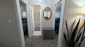 Un pasillo con piso alfombrado que conduce a un baño con ducha, inodoro y lavamanos. Hay dos camas con ropa de cama azul en habitaciones contiguas.