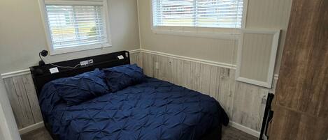 Un dormitorio con una cama, dos ventanas con persianas, un radiador y una puerta.