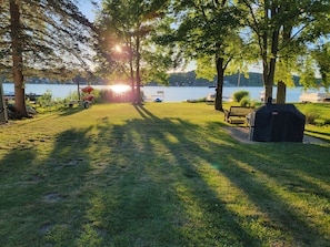 A grassy area with a barbecue grill, a bench, and a view of a lake with boats in the background.