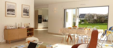 A spacious living room with a glass coffee table, a laptop, and a sofa. There is a dining area with a table set for two and a view of a garden through large windows.