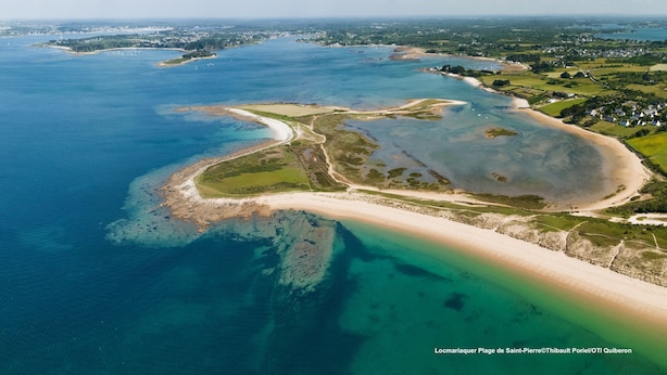 Vue aérienne d’une zone côtière avec une plage, des eaux cristallines et une végétation luxuriante environnante.