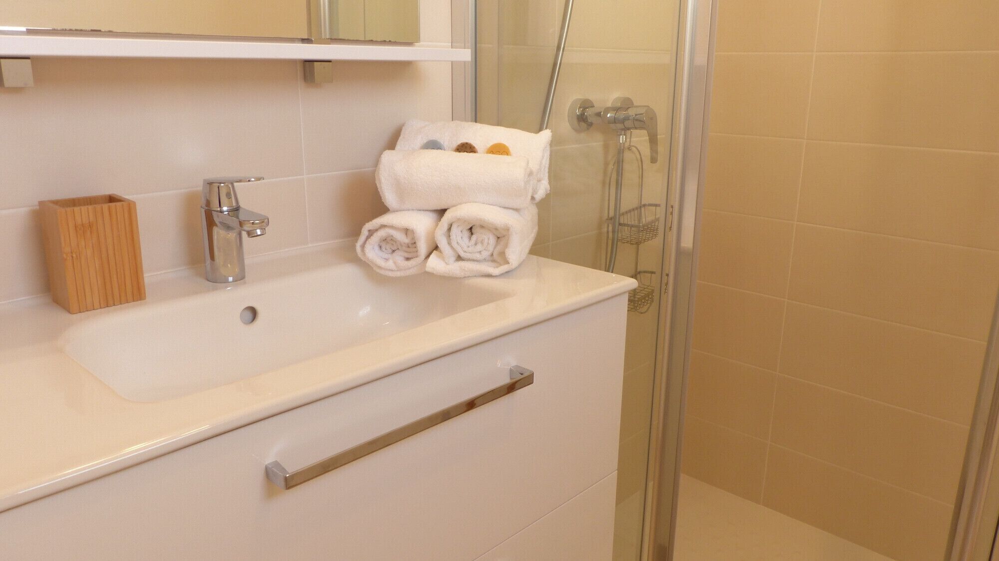 Een badkamer met een wit wastafel, een houten zeepbakje, een handdoekrek met opgevouwen handdoeken en een glazen douchecabine.