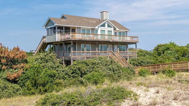 A two-story beachfront house with a balcony, surrounded by vegetation.