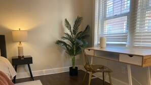Un dormitorio con cama, mesita de noche, lámpara, escritorio, silla, planta en maceta y ventana con persianas.