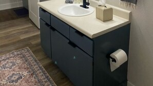 Un baño con un espejo redondo, un lavamanos, un perchero para toallas y un dispensador de papel higiénico.