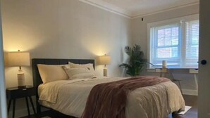 Un dormitorio con una cama, dos mesitas de noche, una lámpara de noche, un ventanal y una planta.