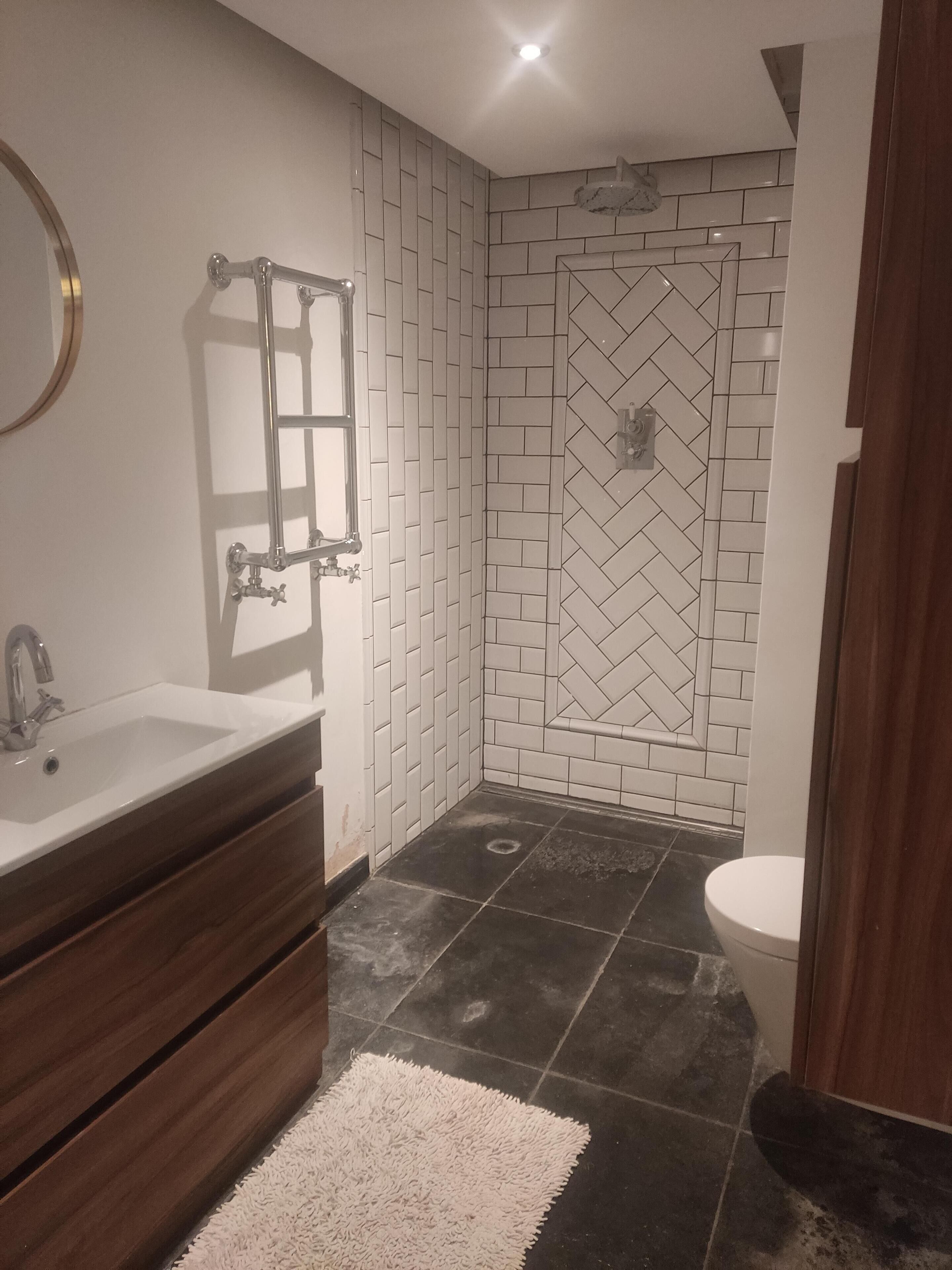 A bathroom with a white sink, wooden vanity, a white rug, a herringbone-patterned shower wall, and a white toilet.