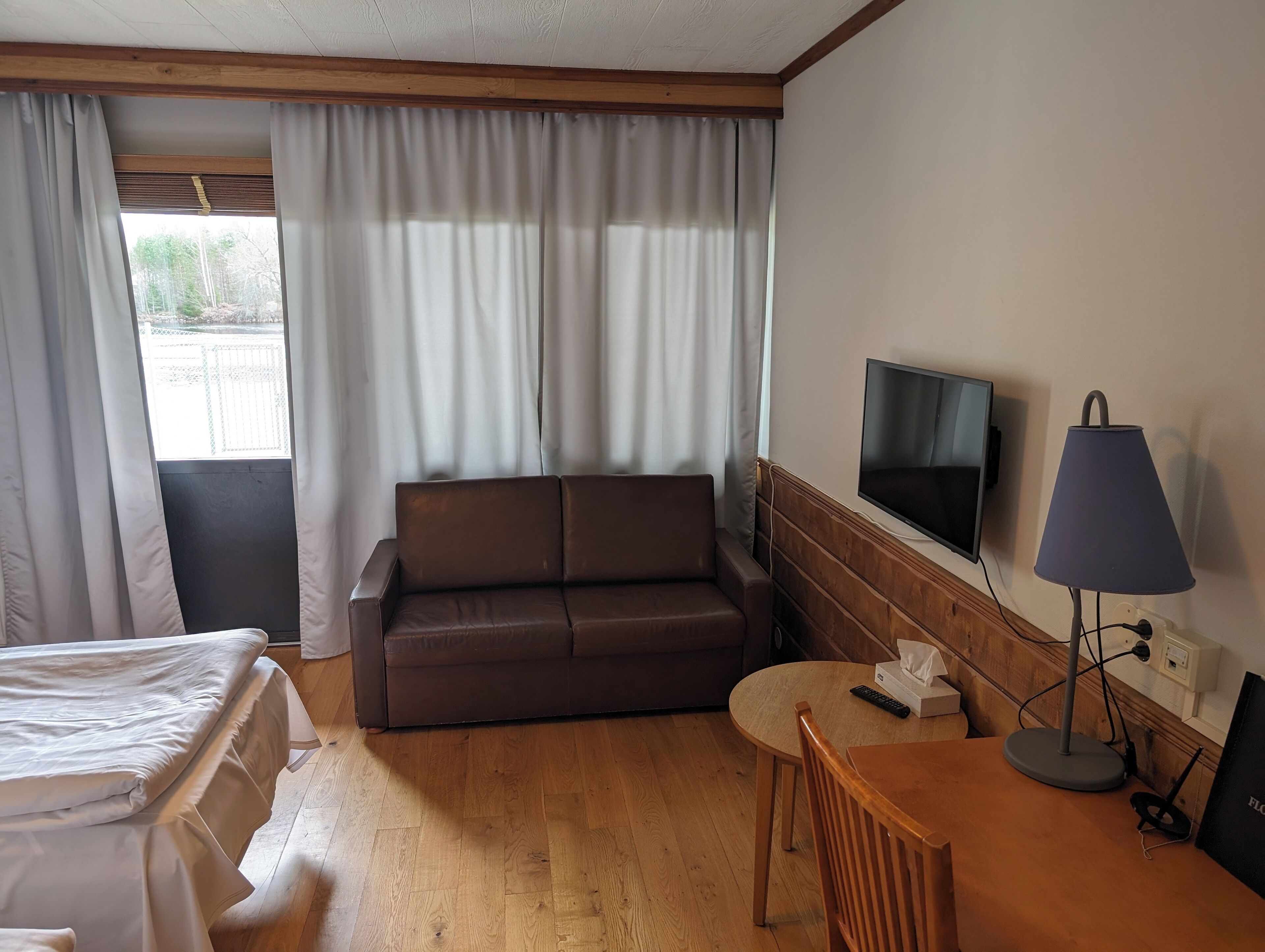 A hotel room with a bed, a brown sofa, a small table, a TV, and a lamp.