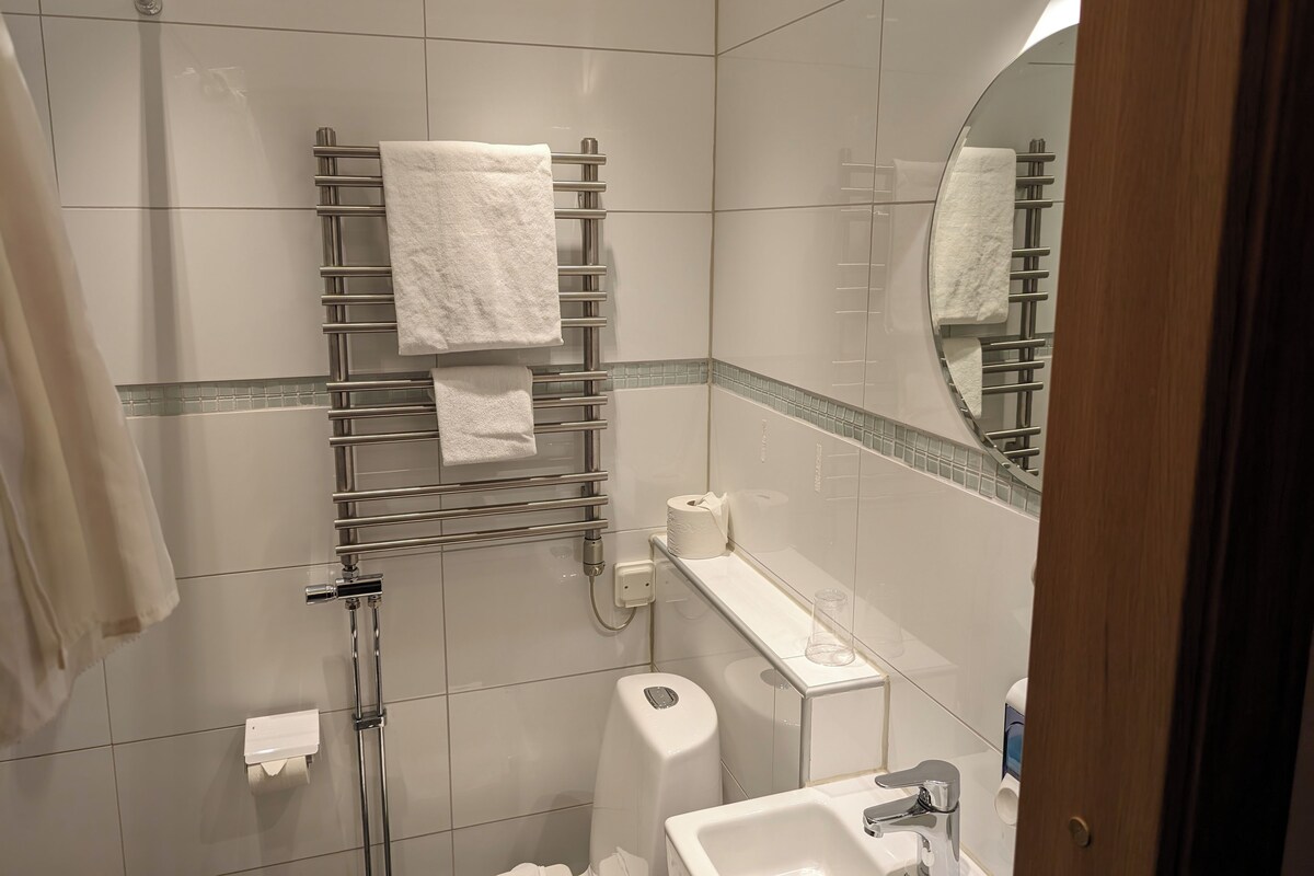 A modern bathroom with a glass shower enclosure, a towel rack with multiple towels, a toilet paper dispenser, and a sink with a mirror above it.