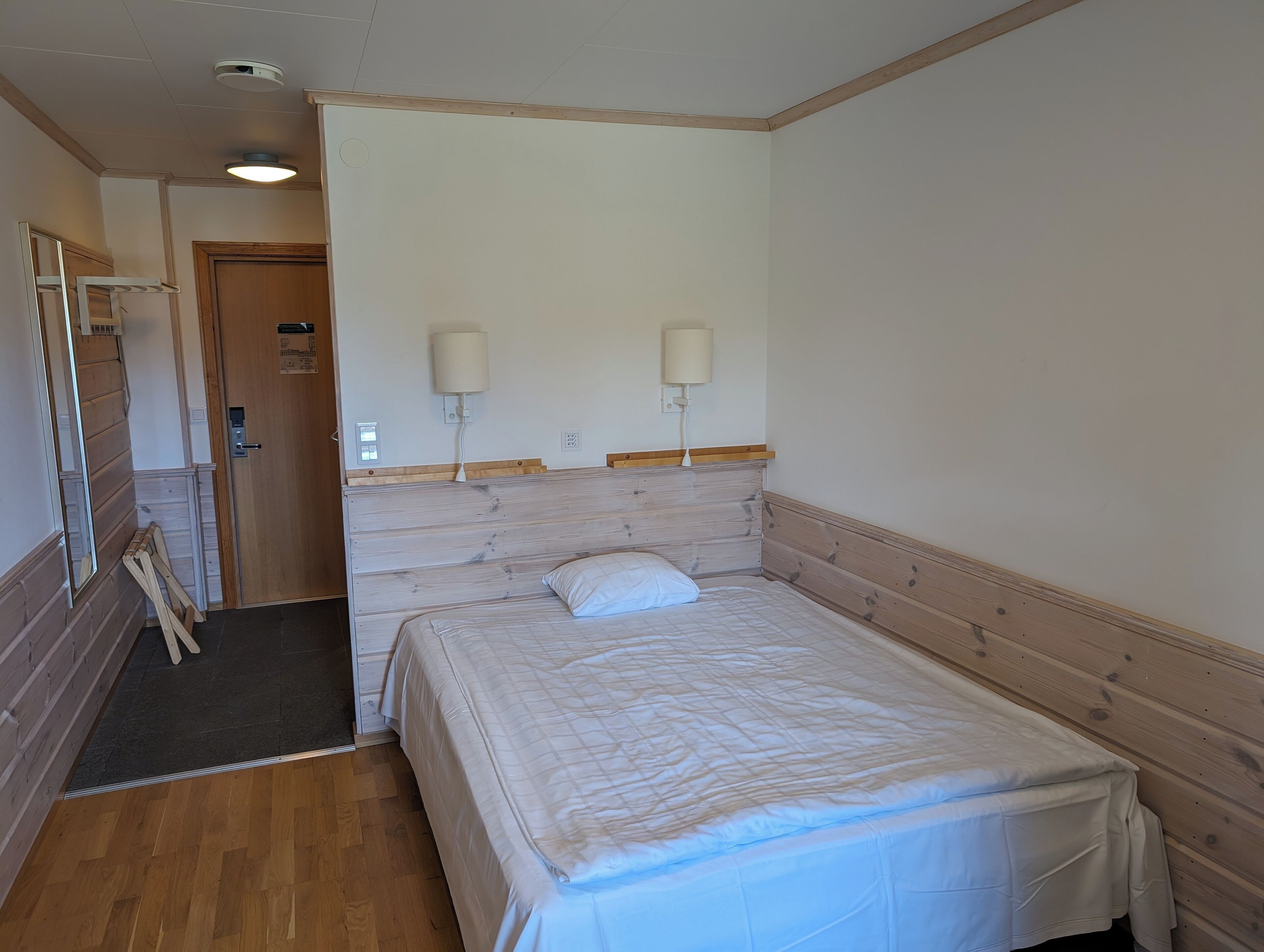 Une chambre d’hôtel avec une tête de lit en bois, un lit avec une literie blanche, deux lampes fixées au mur, une porte avec une pancarte et un plancher en bois.