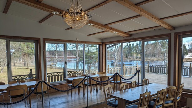 A spacious dining area with wooden tables and chairs, large windows offering views of a lake and surrounding trees, and a chandelier hanging from the ceiling. - Värdshuset Flottaren (Vansbro)