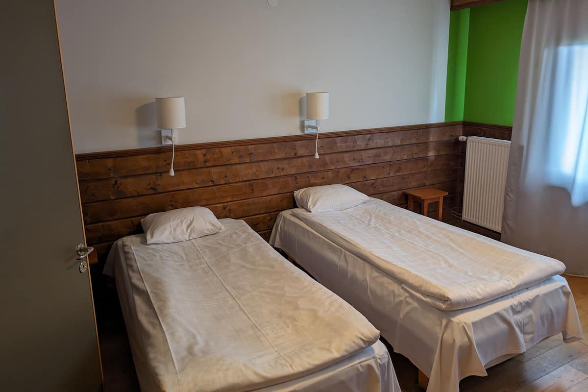 Une chambre avec deux lits simples, une tête de lit en bois, des murs blancs, deux lampes de chevet, une petite table de chevet en bois et un radiateur.
