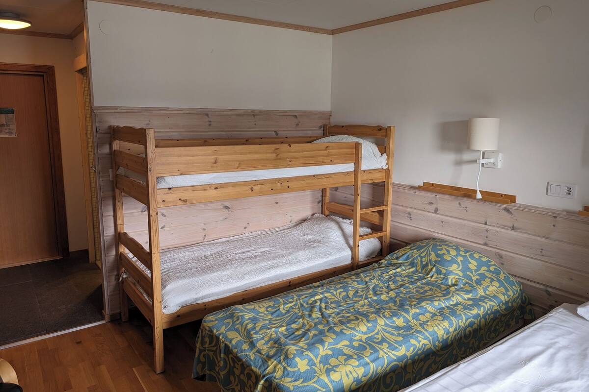 Un lit superposé avec une structure en bois, un lit inférieur avec un matelas blanc et un lit supérieur également avec un matelas blanc. Il y a une table de chevet avec une lampe, une couvre-lit à motifs et une porte sur le côté gauche.