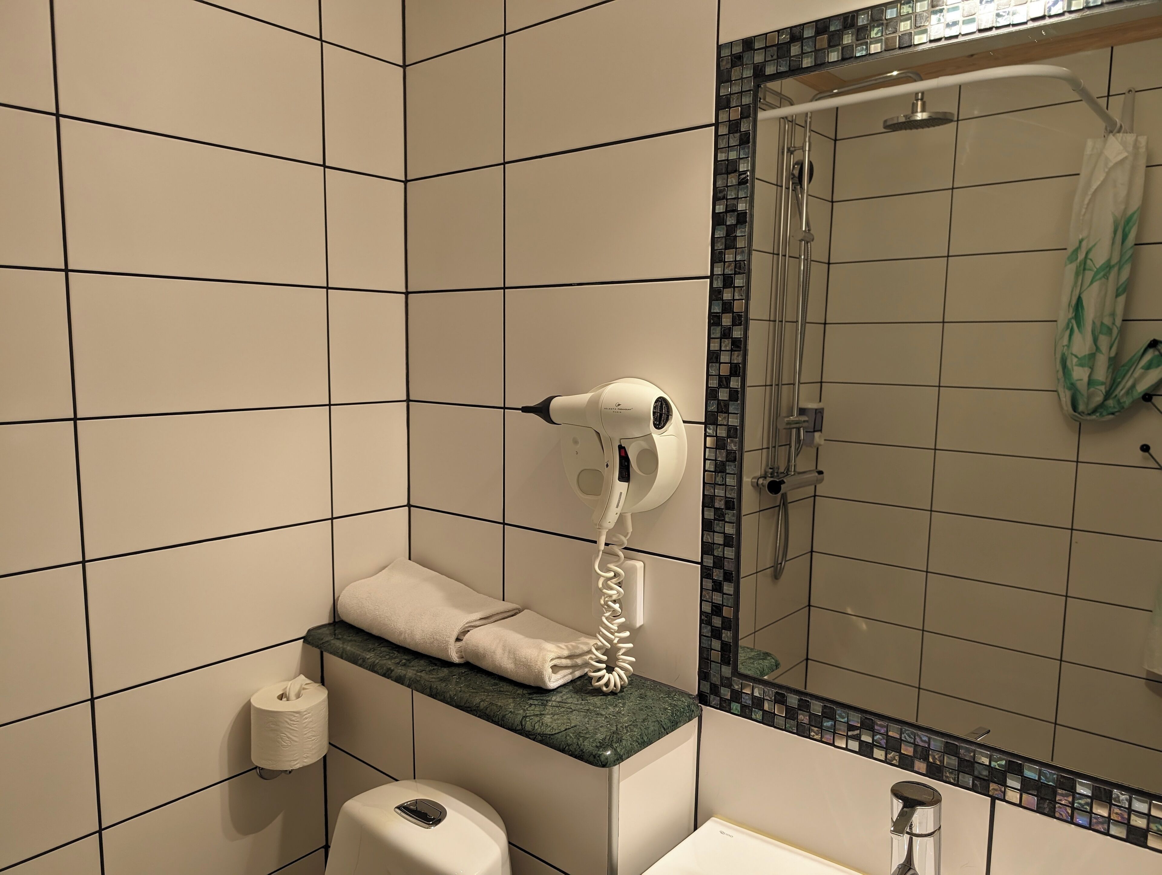 Un baño con toallero, secamanos, dispensador de papel higiénico y zona de ducha con pared de azulejos.