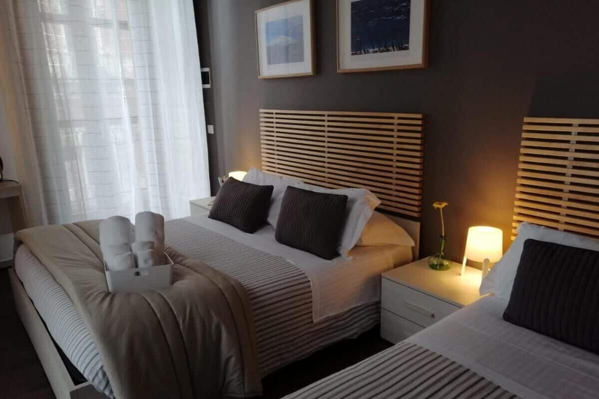 Habitación de hotel con dos camas, una mesita de noche con lámpara y una ventana con cortinas traslúcidas.