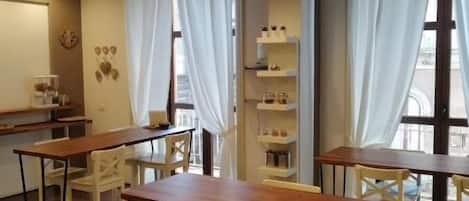 A dining area with wooden tables and chairs, a hanging light fixture, and a view of the street through large windows.