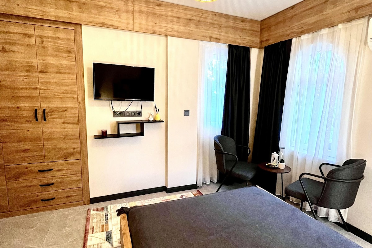 A modern bedroom with a large bed, a flat-screen TV mounted on the wall, a wooden wardrobe, and a small table with a chair.