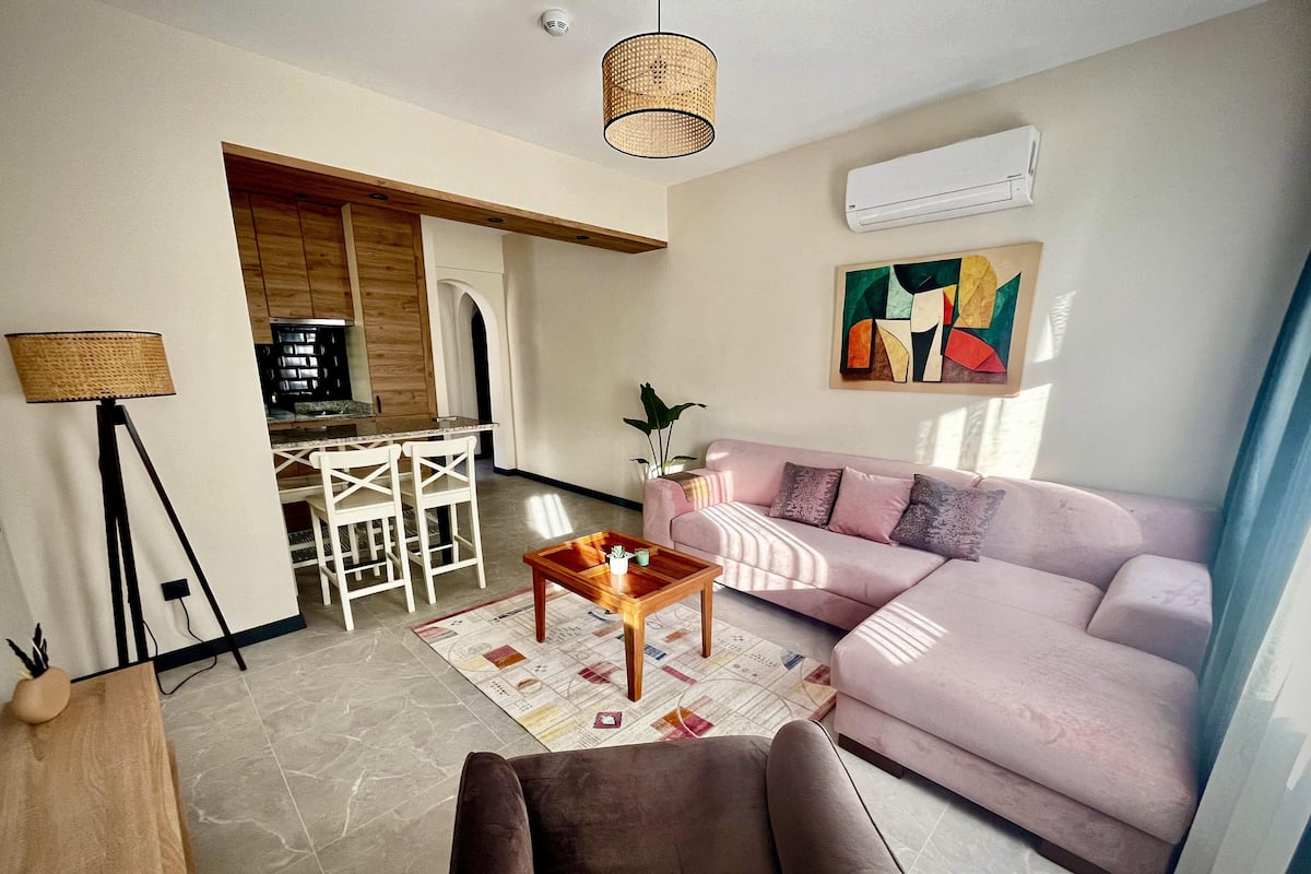 A modern living room with a pink sofa, a wooden coffee table, and a dining area with white chairs.