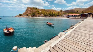 A coastal scene with a wooden pier extending into calm waters, a small boat anchored nearby, and a hill with buildings and palm trees in the background. - Kay Alice - Beachfront Villa (Le Carbet)