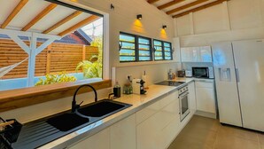A modern kitchen with white cabinetry, a built-in oven, a microwave, and a refrigerator. There are wooden beams on the ceiling and pendant lights above the counter. - Kay Alice - Beachfront Villa (Le Carbet)