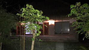 A nighttime view of a building with a thatched roof, illuminated windows, and a small water feature in the foreground. - Melody (Jerowaru)
