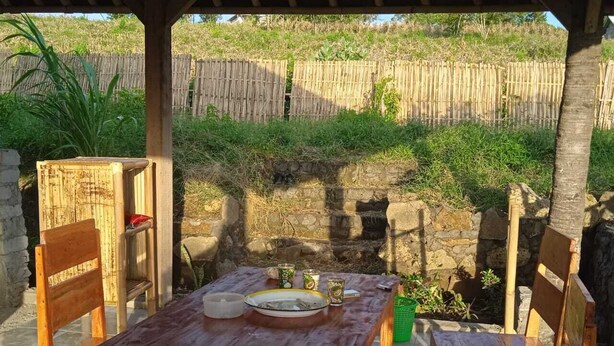 An outdoor wooden table and chairs set under a wooden pergola, with a stone wall and bamboo fence in the background. - Melody (Jerowaru)
