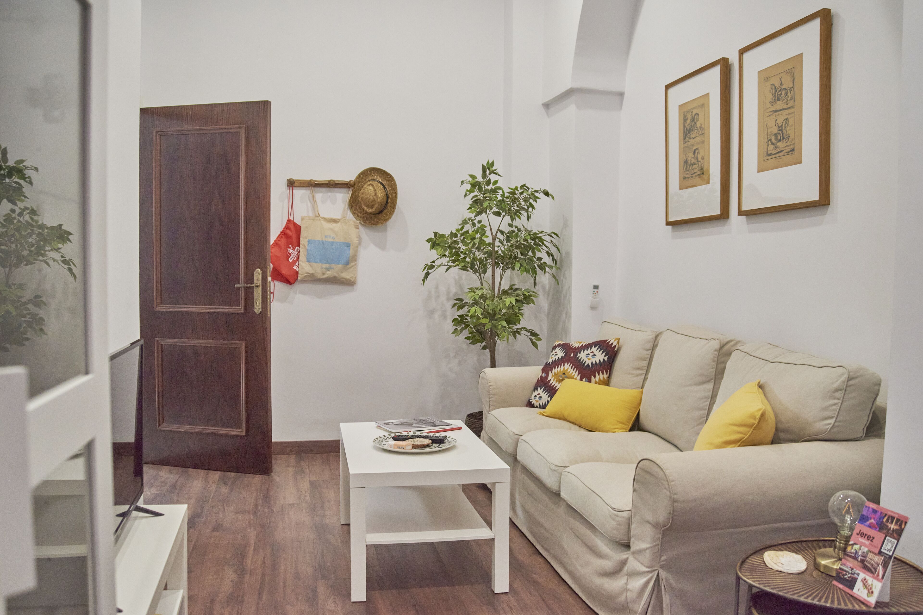 Ein Wohnzimmer mit einem beigefarbenen Sofa, einem kleinen weißen Tisch, einer Topfpflanze und zwei gerahmten Kunstwerken an der Wand.