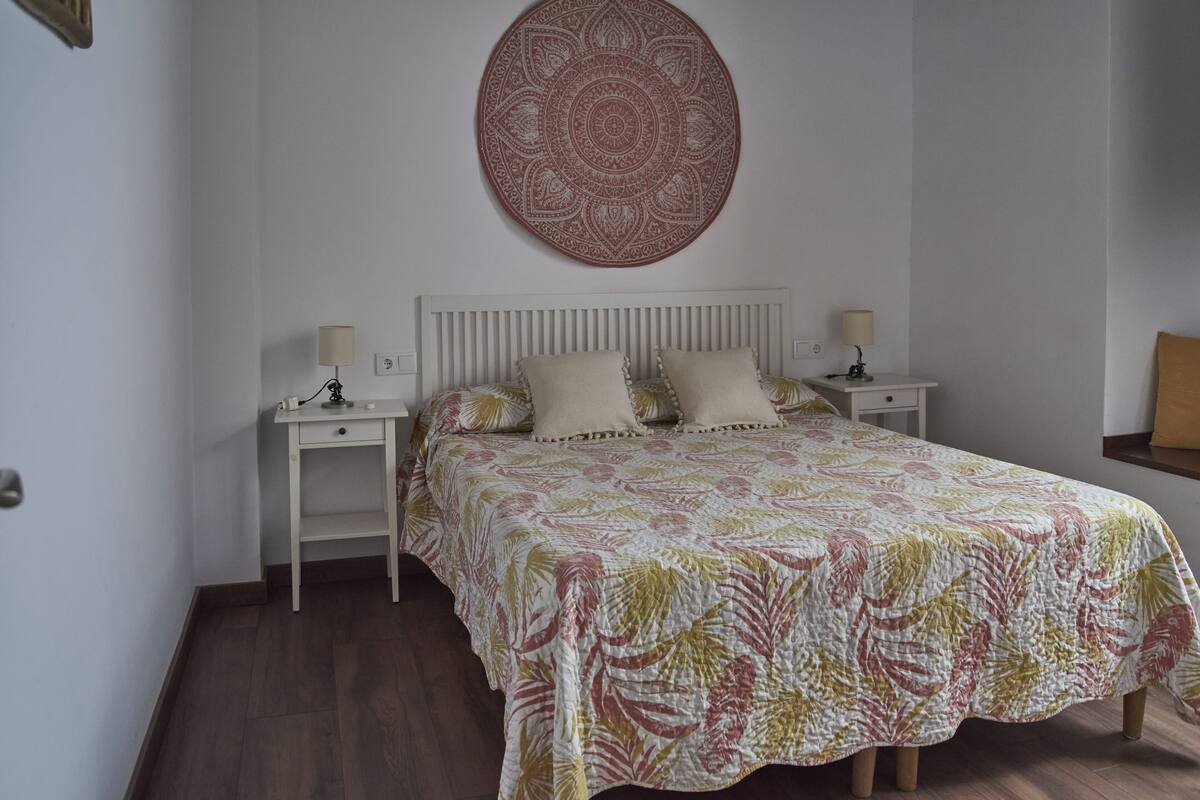 A bedroom with a bed, two bedside tables, a wall hanging, and a nightstand.