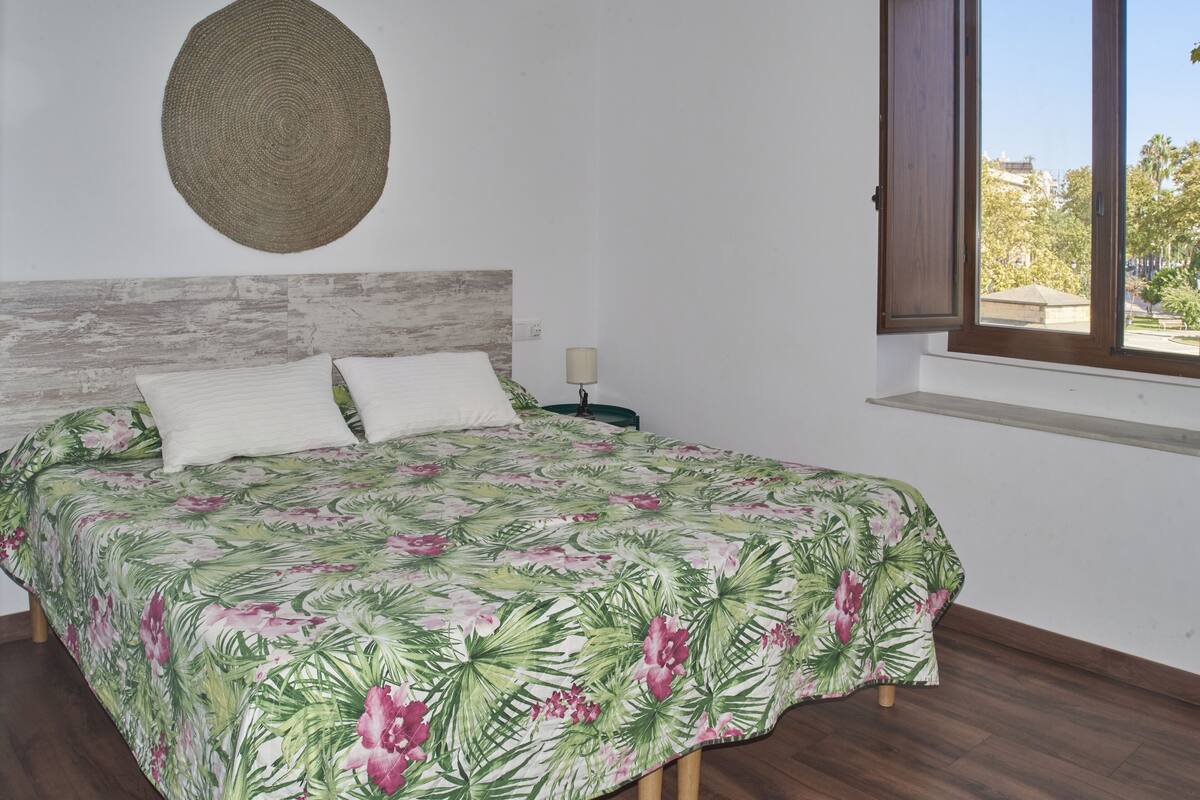A bed with a floral bedspread, a woven wall hanging, and a window with a view of trees and buildings.