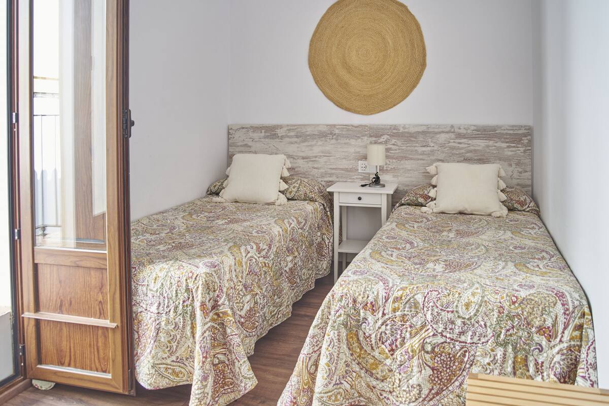 A bedroom with two beds, a wooden door, a nightstand with a lamp, and a woven wall hanging.
