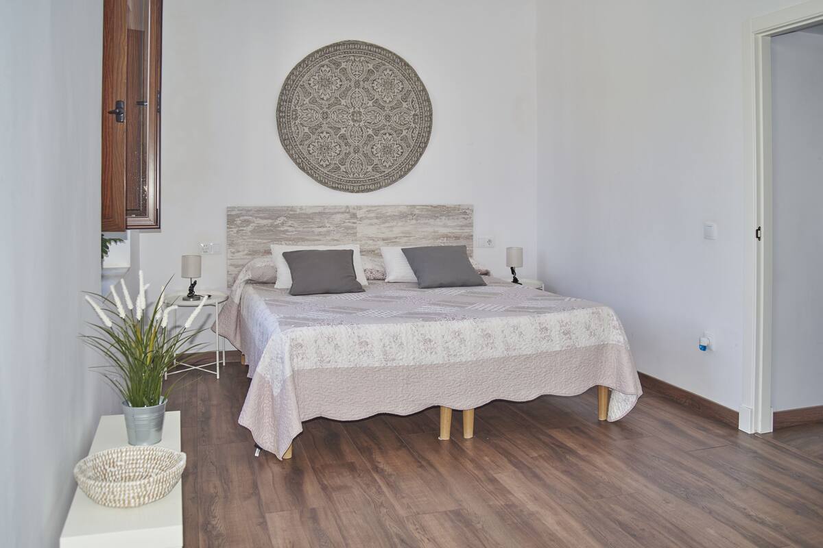 A bedroom with a bed, a round wall hanging, a bedside table with a lamp, a potted plant, and a woven basket.