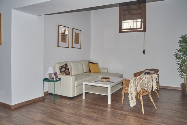 A living room with a sofa, coffee table, side table, wicker chair, and a potted plant. - Apartamentos Mamelón (Jerez de la Frontera)