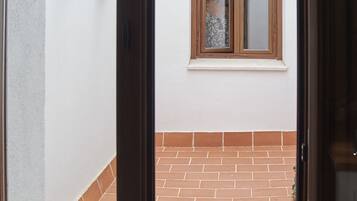 Vista a través de una puerta a un patio con piso de ladrillo, una ventana con contraventanas de madera y plantas en macetas en la pared.