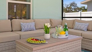 A cozy outdoor seating area with a wicker coffee table, a sofa with cushions, a vase of white flowers, and a tray with fruits and drinks.