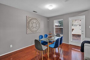 A dining area with a glass table, blue and gray chairs, a decorative wall piece, and a door with a view. - Frisco Urban Escape (Frisco)