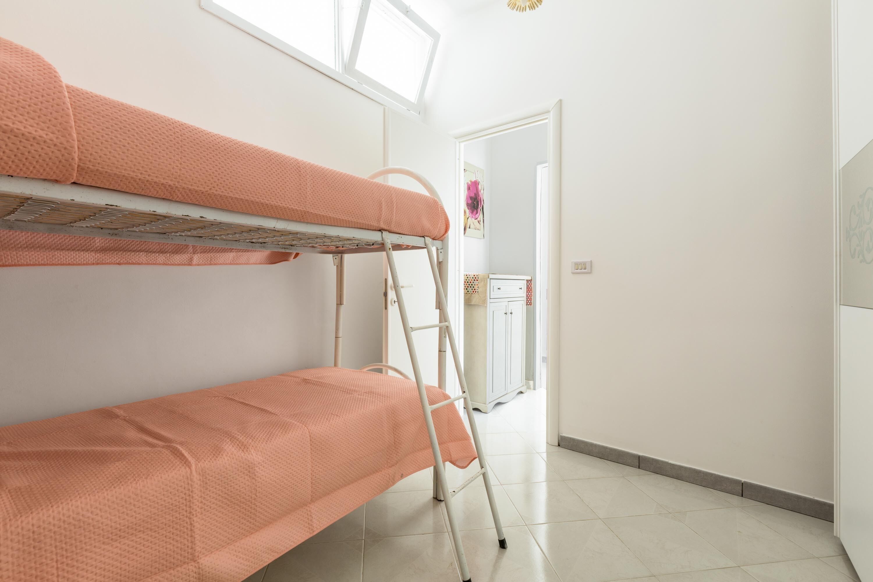 A bunk bed with a ladder, a white door, and a small cabinet in a room with white walls and tiled flooring.