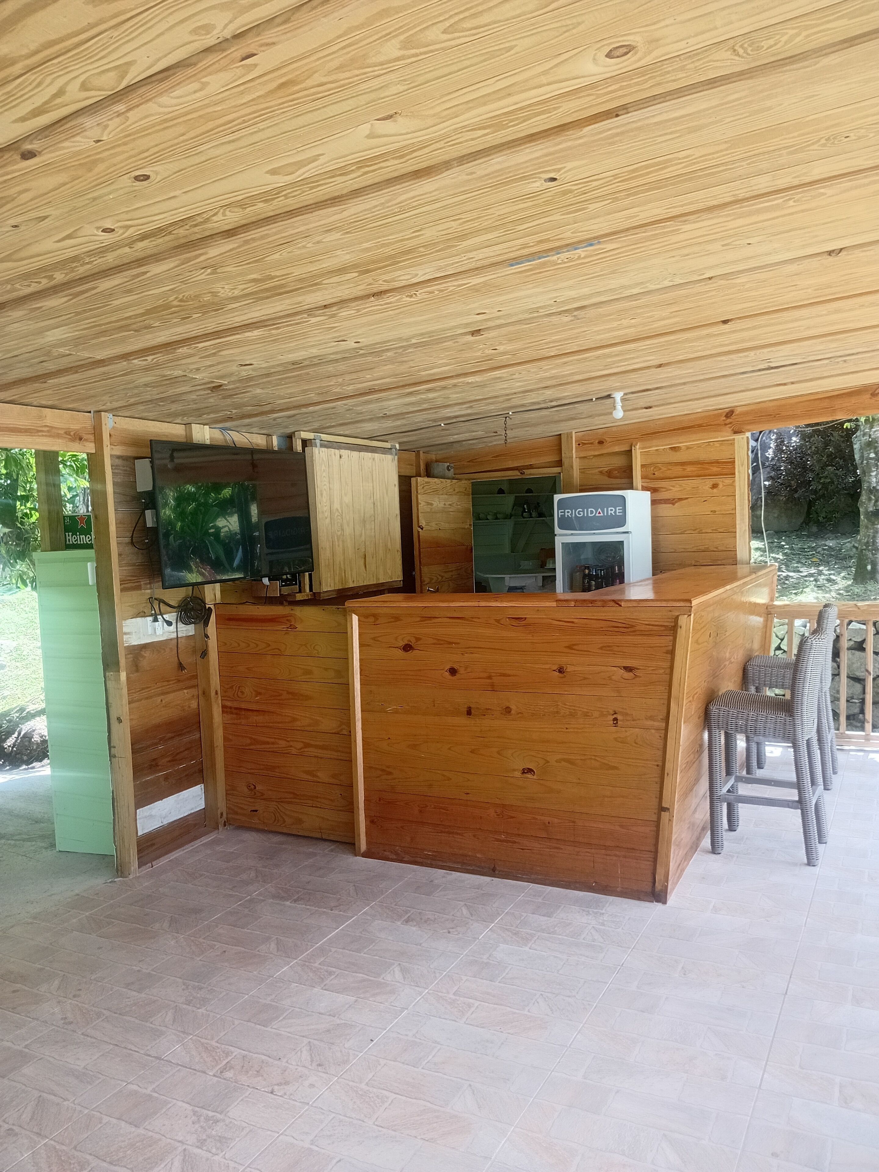 A wooden kitchen with a refrigerator, a television, and a bar area with wicker chairs.
