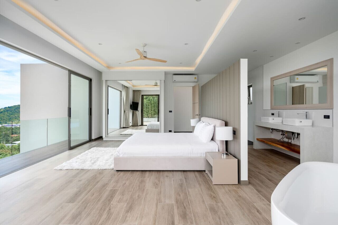 A modern bedroom with a large bed, a walk-in closet, a bathroom with a bathtub, and a balcony with a city view.