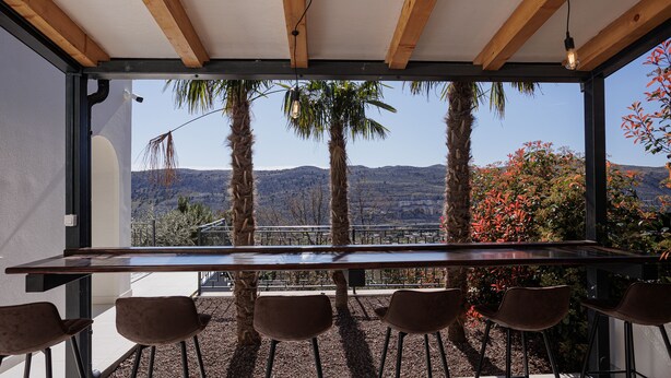 A modern outdoor dining area with a long table and six chairs, overlooking a scenic landscape with mountains and trees.