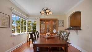 A dining room with a wooden table and chairs, a chandelier, a window with blinds, and a kitchen area with cabinets. - Home away from home in the heart of charming Winter Park! (Winter Park)