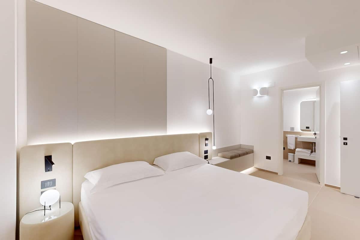 A modern bedroom with a large bed, bedside tables, a built-in bench, and a bathroom with a mirror and sink.